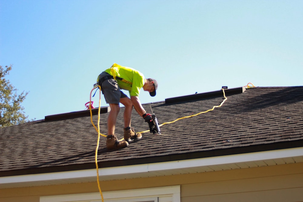 Roofer in safety harness drilling into a shingle roof on a sunny day